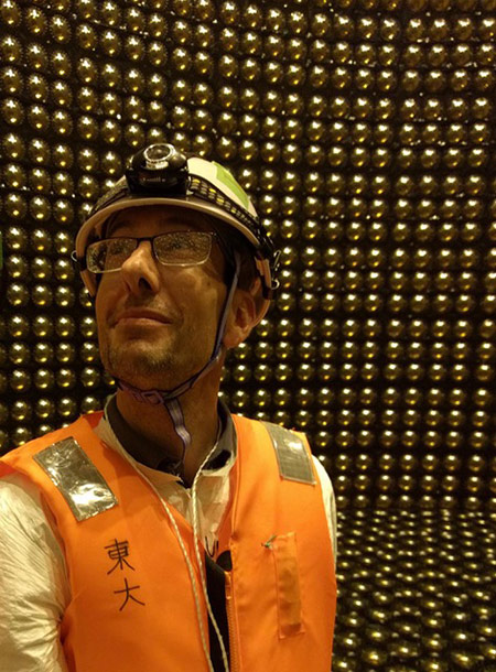 professor in a hard hat and safety vest stands in a large detector array.
