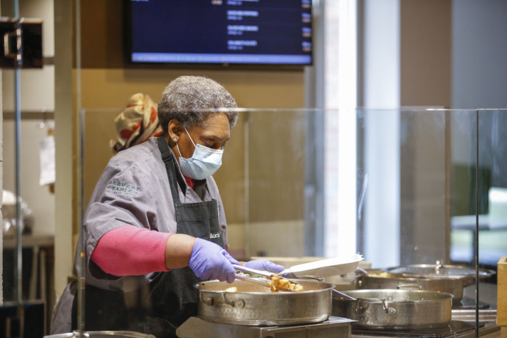 Gloria Jackson wears a face mask and uses cooking tongs to fill a to-go lunch container.