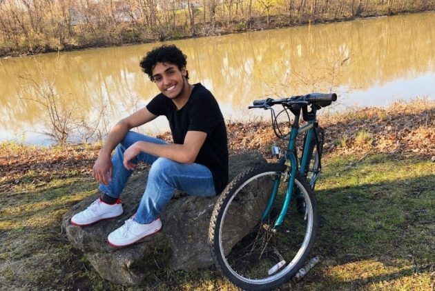 Student sits by river with bicycle.