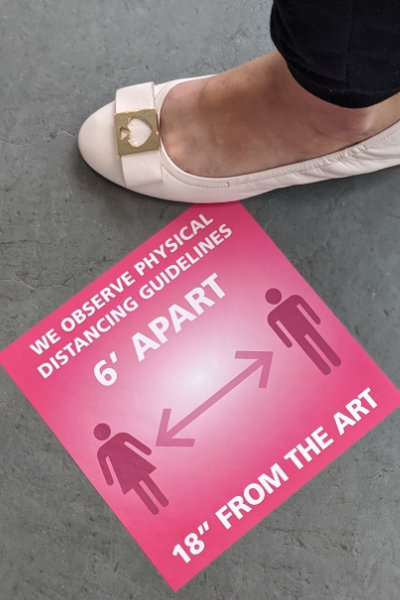 Foot wearing white shoe next to bright pink floor decal at MAG asking people to stay six feet from each other and 18 inches from the art.