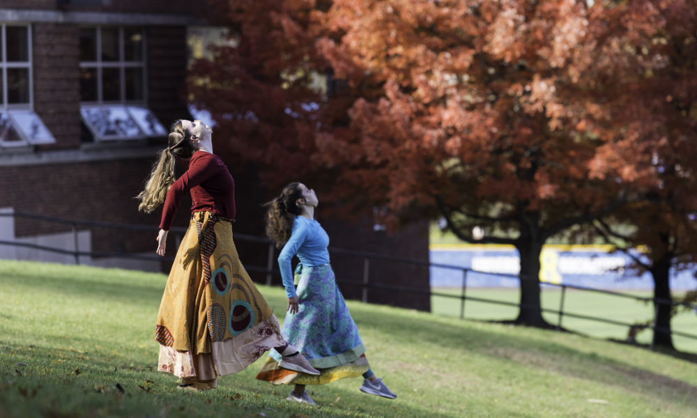 Two masked women in colorful garb dance down a hill on campus.