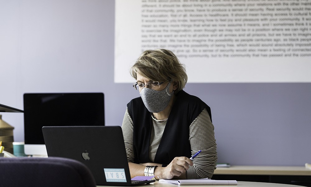 professor sitting at their desk, wearing a mask and working on a laptop.