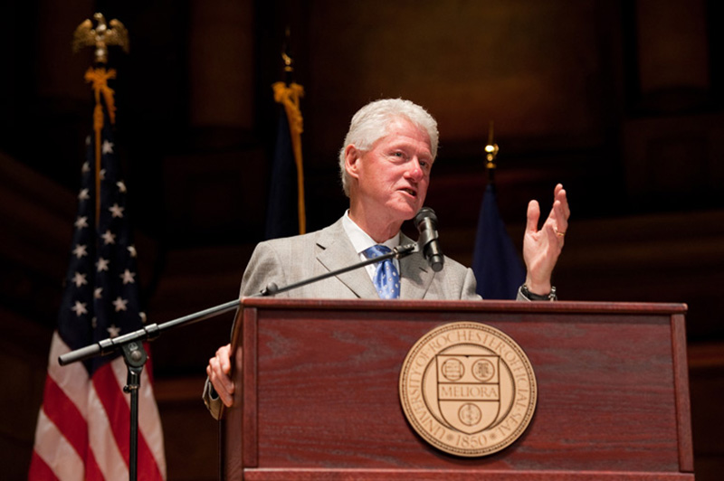 archival photo of Bill Clinton speaking behind a podium with a University of Rochester seal. 