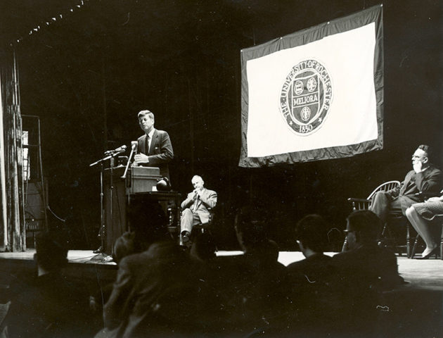 archival photo of JFK speaking at a podium in front of a University of Rochester flag.