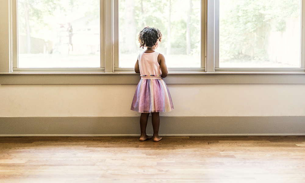little Black girl seen from behind looking out a bay window.