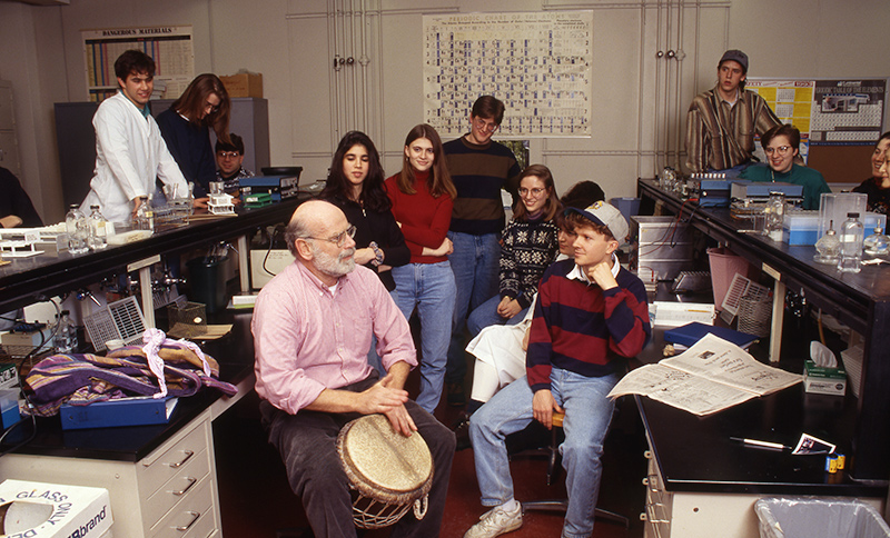 archival photo of Professor Hattman in his lab, surrounded by a group of students, as he plays an African drum.