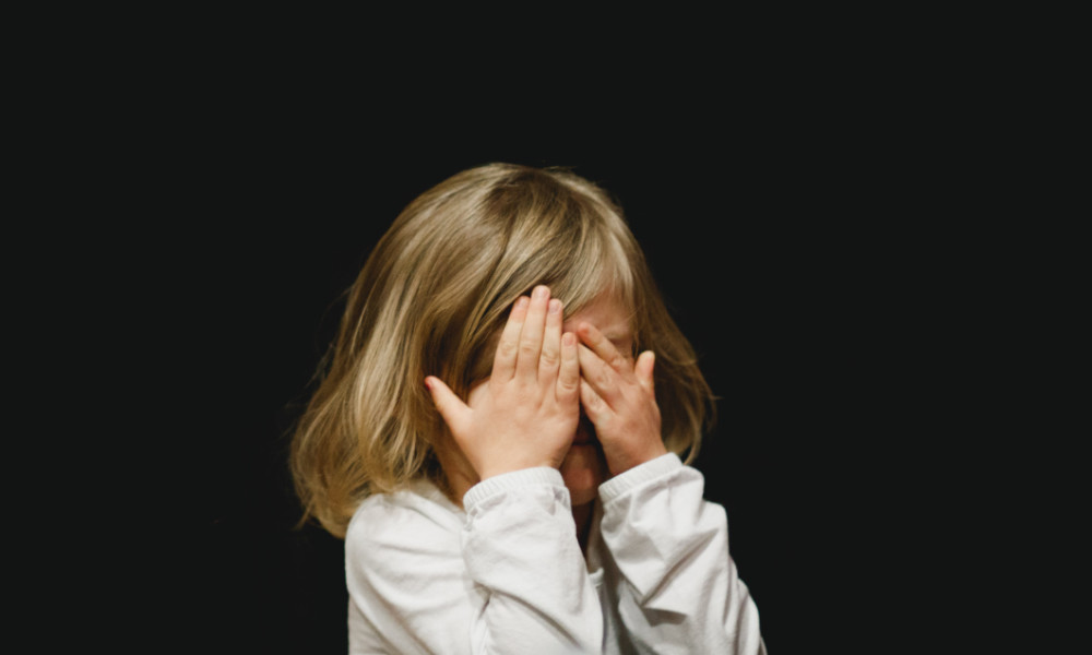 Young blonde girl against black background wears white long sleeve shirt and covers her eyes.