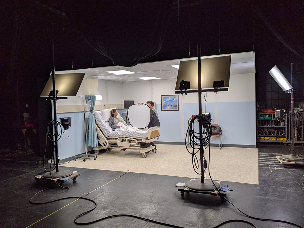two actors on a set, one in a hospital bed and the other sitting on the side, holding a lighting reflector.