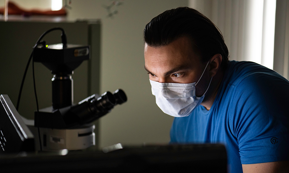 Jeffrey Beard wears a mask and blue shirt while leaning in near a microscope.