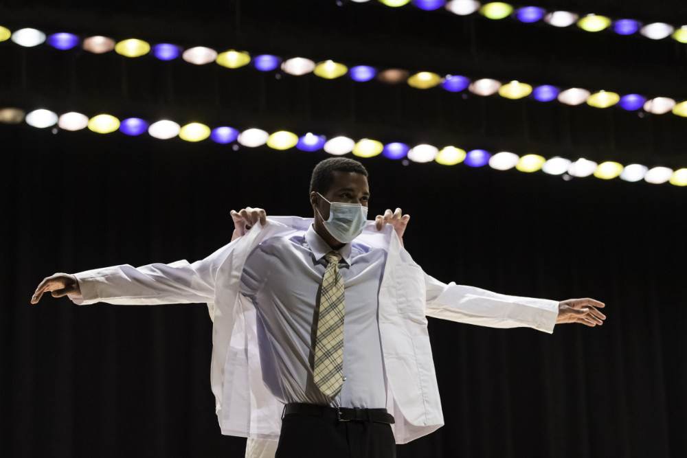 Student wearing slacks, shirt, tie, and mask has someone help him with his white coat.