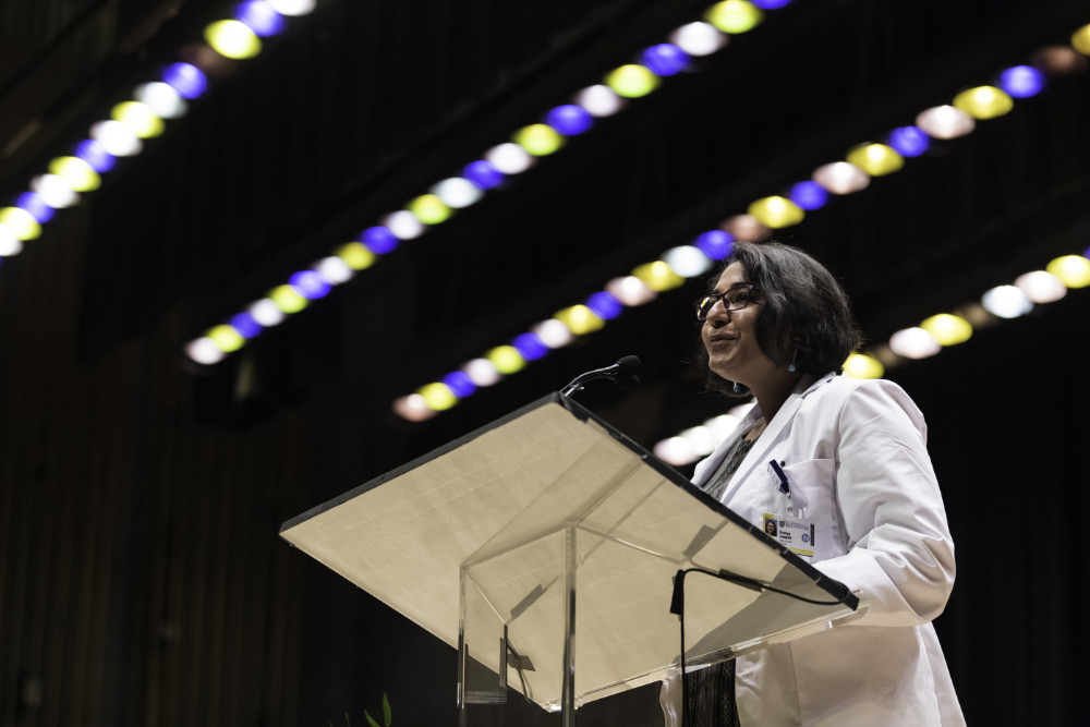 Ramya Sampath stands at a podium in a white coat with blue, yellow, and white lights shining overhead.