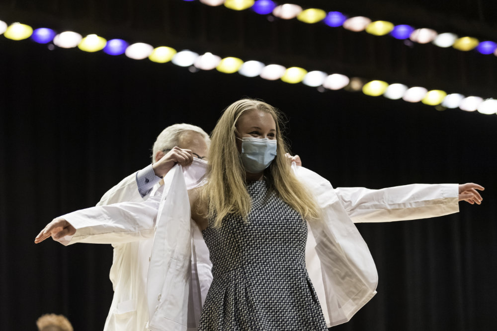 Female student wearing a masks gets help putting on her white coat. 