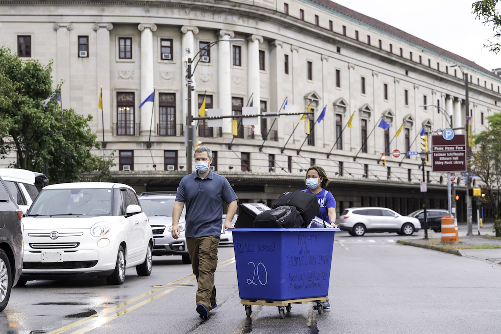 parents push a wheeled blue bin with their son's belongings with Eastman Theatre in the background