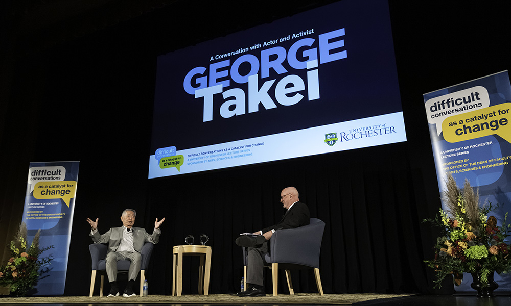 George Takei and Dean Donald Hall seated on stage under a screen that reads George Takei.