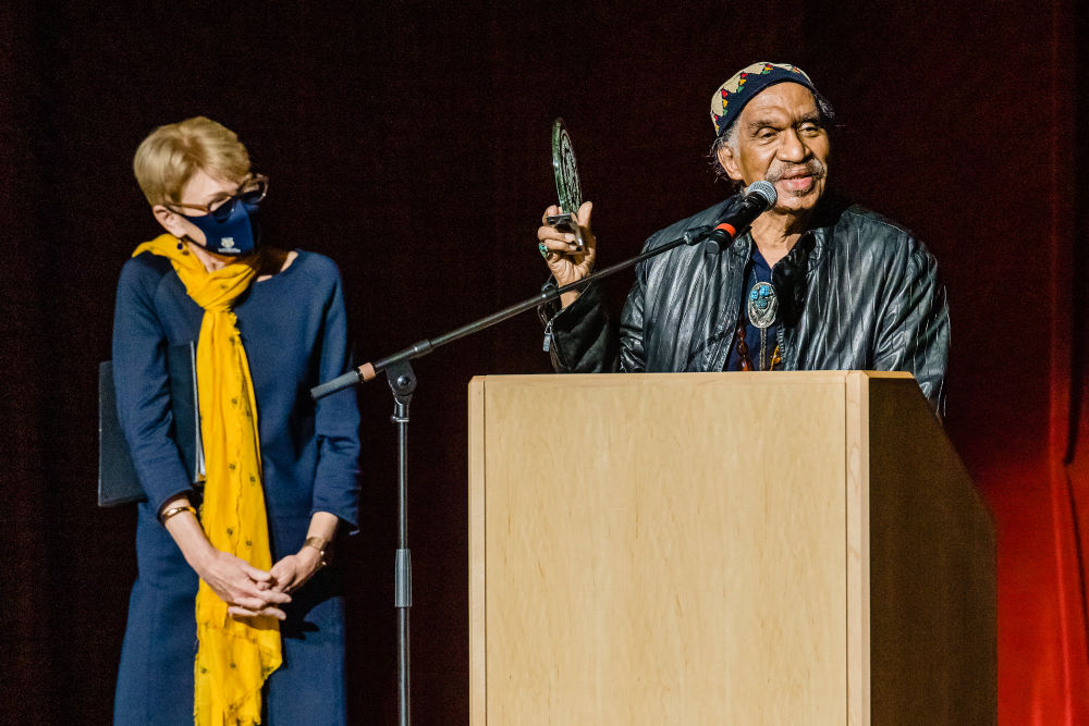 Sarah Mangelsdorf in a blue dress, yellow scarf, and mask looks at Garth Fagan, who is standing next to her at a podium holding an award.