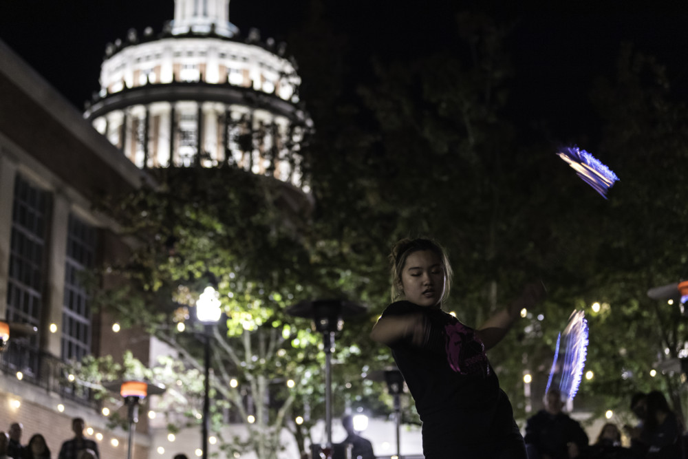 Student juggles fire props at night with onlookers to the side and Rush Rhees Tower lit up in the background.