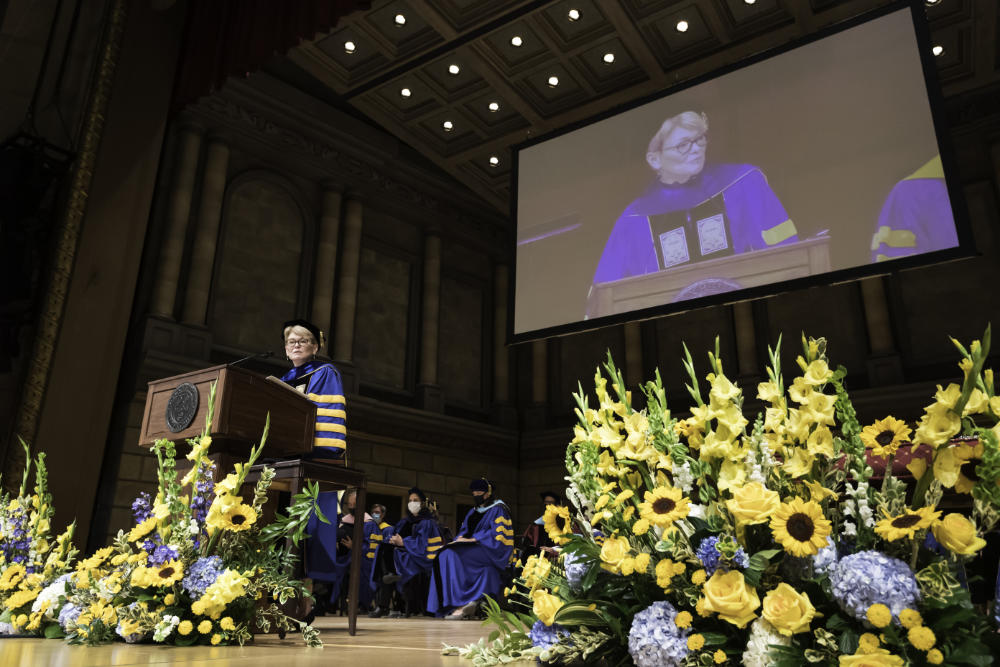 Regalia-clad Sarah Mangelsdorf stands at a podium on a flower-covered stage while her image projects on a big screen behind her for Meliora 2021 commencement.
