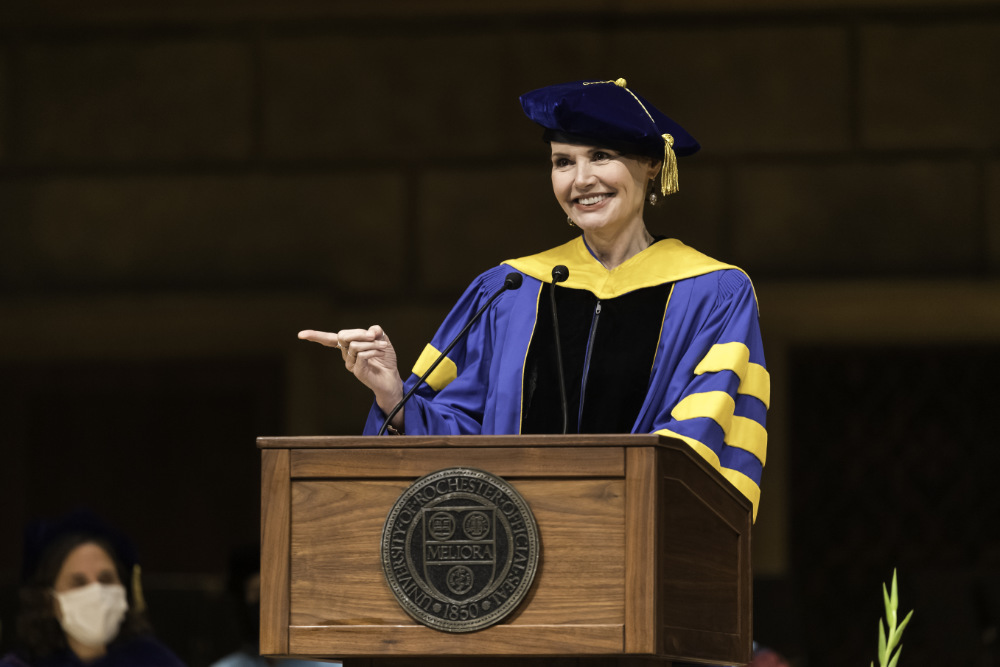 Geena Davis wears University of Rochester regalia while standing at a podium, smiling, and pointing at the audience.