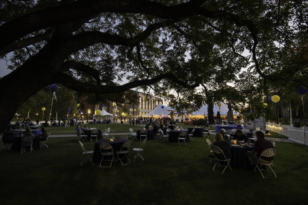 Meliora Village tables, tents, decorations, and guests at dusk with tree branches in the foreground.