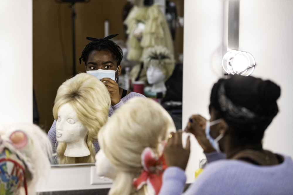 Student in mask works on a costume wig while looking in the mirror.