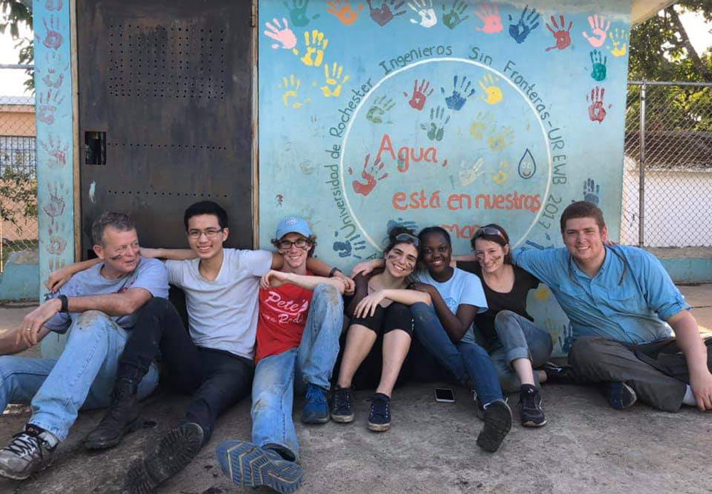 large group of students sitting on the ground in front of a building decorated with colorful painted handprints.