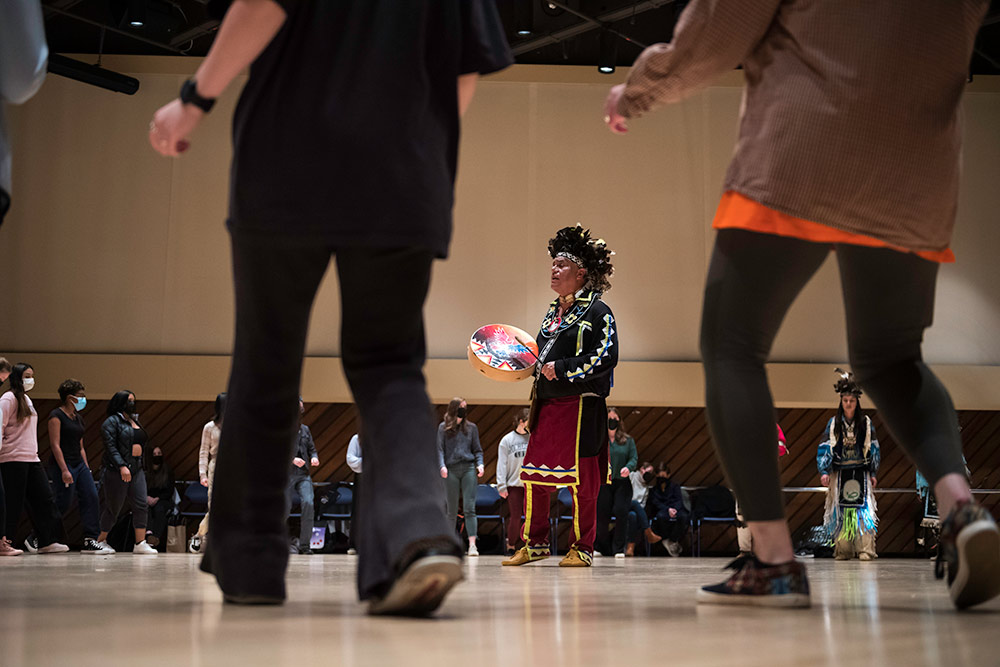 A dancer in traditional Native American dress stands in the center of a circle of people, playing a drum. We view him through the legs of the people standing around him in a circle.