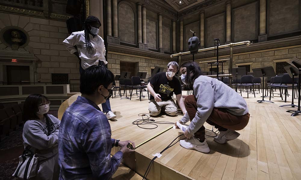 Two people hold measuring tape on Kodak Hall stage while two others look on. 