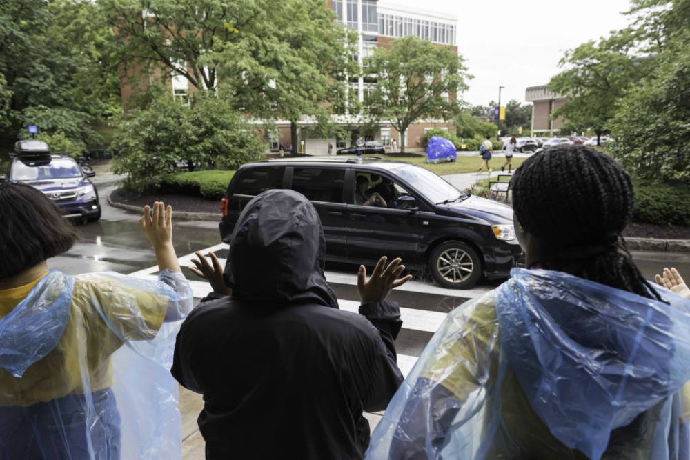 Seen from behind, three people wave at and welcome cars of new students.