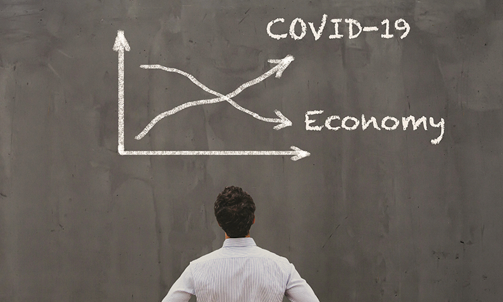 man looks at chalkboard that shows a line graph with trending upward for Covid-19 and down for economy.