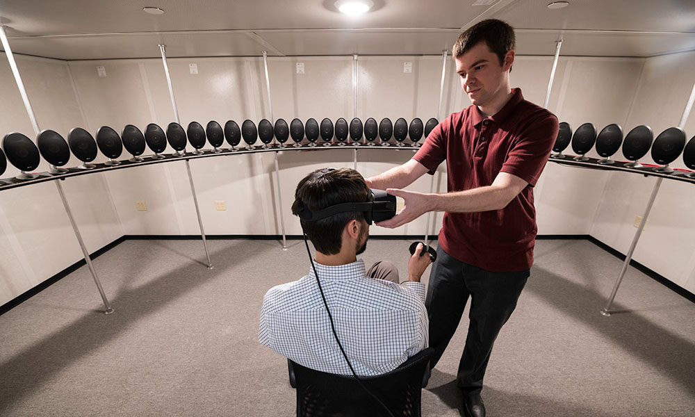 person sitting in a chair wearing a virtual reality headset and surrounded by speakers as another person adjusts the headset.