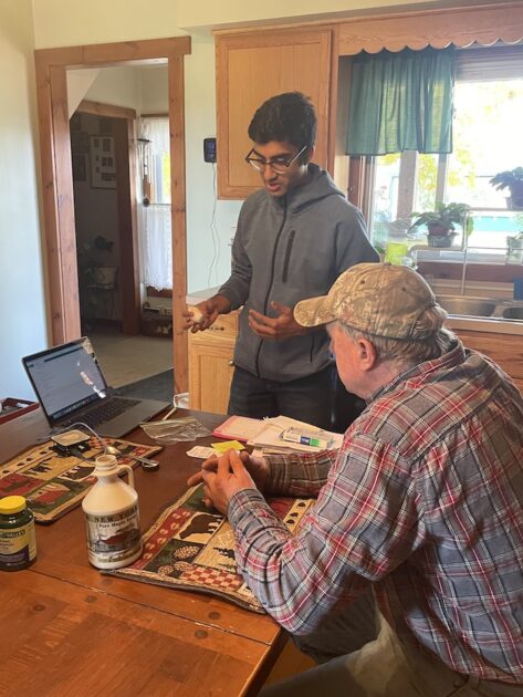 Student and man talk around a kitchen table that has a jar of maple syrup and a laptop on it.