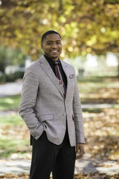 Cory Hunter in a suit and with his hand in his pocket smiles for a photo in autumn on the quad.