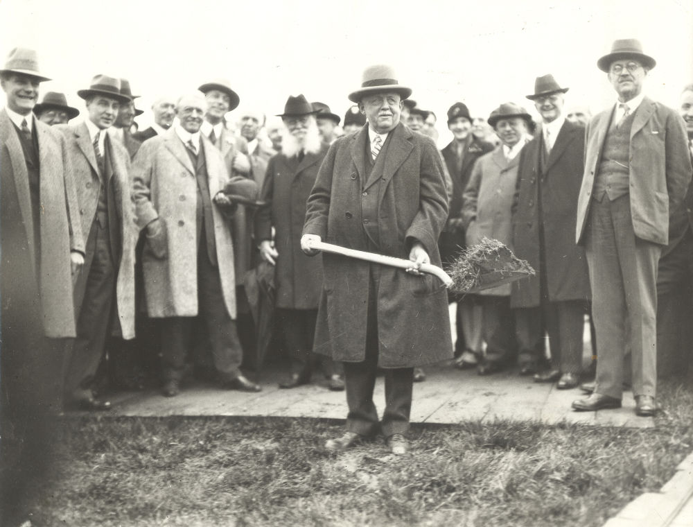 Black-and-white archival photos of men in suits and coats surrounding one such man holding a shovel.