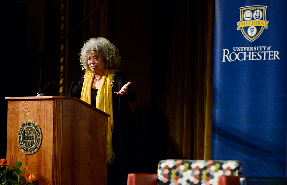 Angela Davis gestures while speaking from behind the podium, with the University of Rochester seal projected in the background.