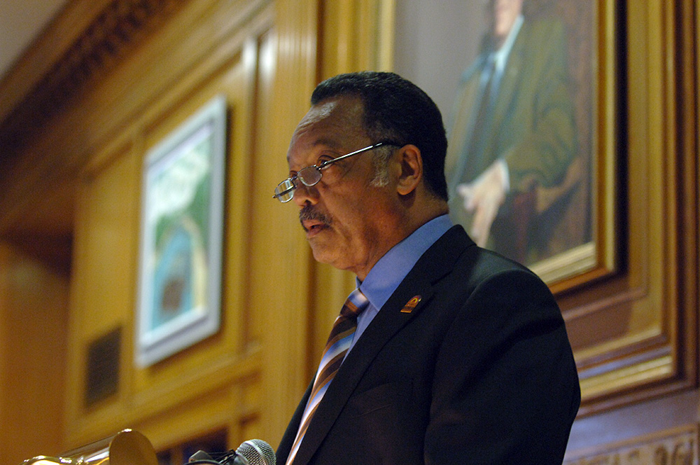 Jesse Jackson, speaking from behind the podium with the wood paneling and portraits of the Welles Brown Room behind him.