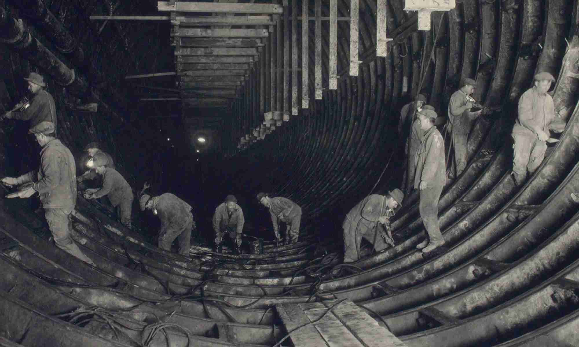 Black-and-white historic photo shows workers cleaning, caulking, pointing, and making the iron watertight invert of New Jersey's South Tunnel east to illustrate biased state spending at the district level.