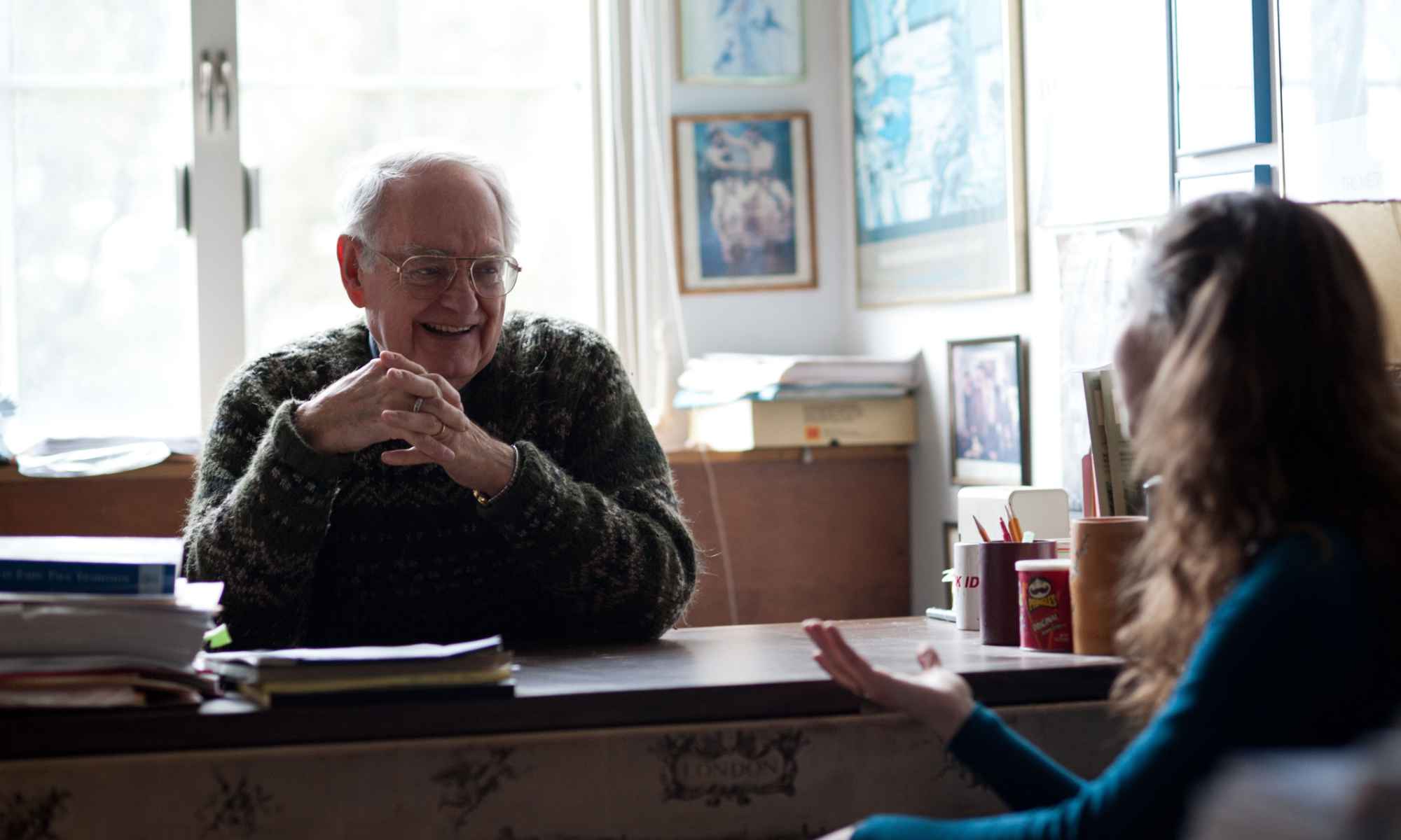 Russell Peck at his desk smiling while talking to a student, who is seen from behind.
