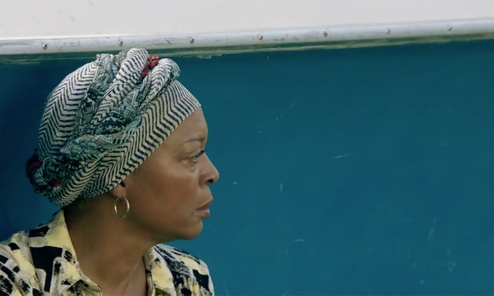 Screenshot of Black actress Tina Lifford, wearing a colorful head wrap and standing in profile against a bright blue wall.