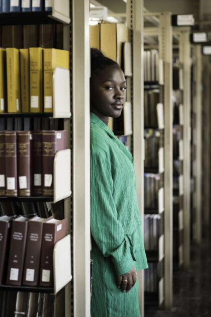 Fatou Jobe, who is translating Wolof fables into English, seen in bright green stands partially hidden by library stacks of books.