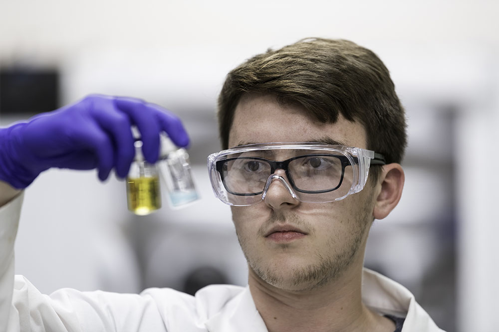 Student in lab goggles, gloves, and coat holds up vial of semiconducting fluid.