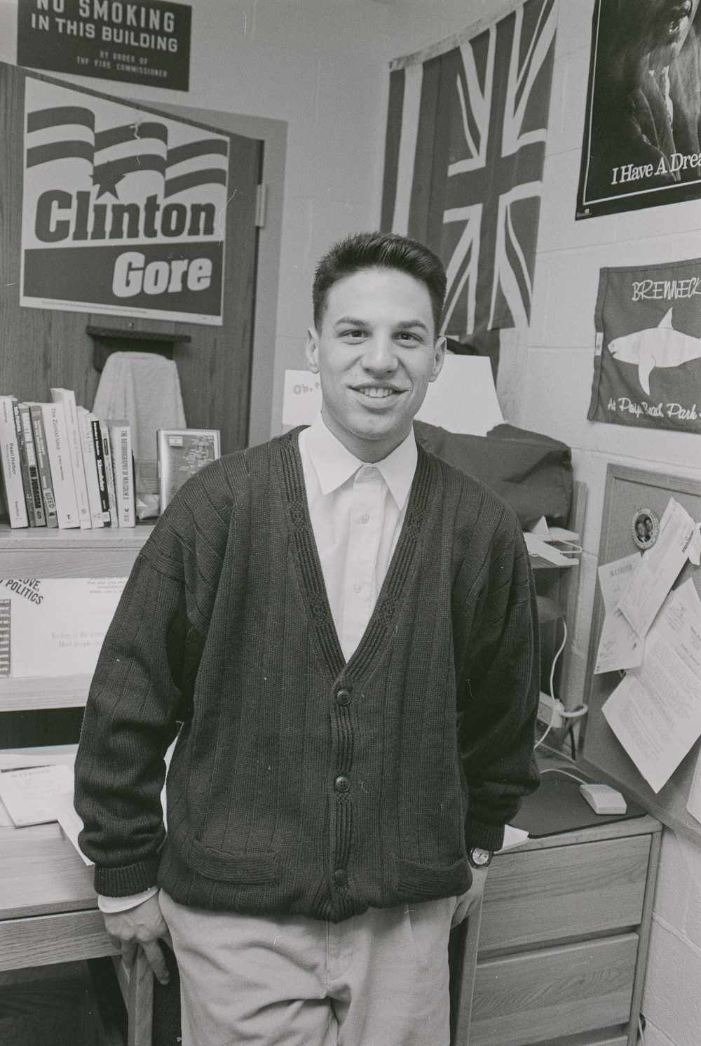Black and white archival photo of Josh Shapiro as an undergraduate in his dorm room with a Clinton/Gore sign, UK flag, and other posters and artwork in the background.