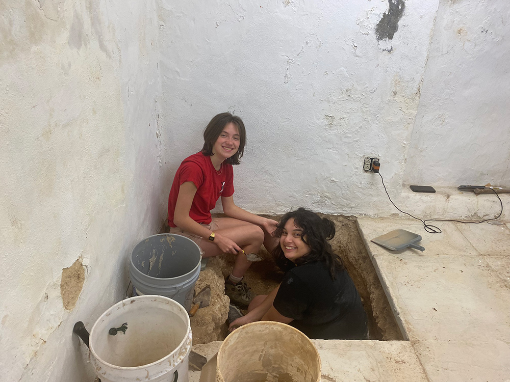Two students digging in a square hole in a corner of a whitewashed structure.
