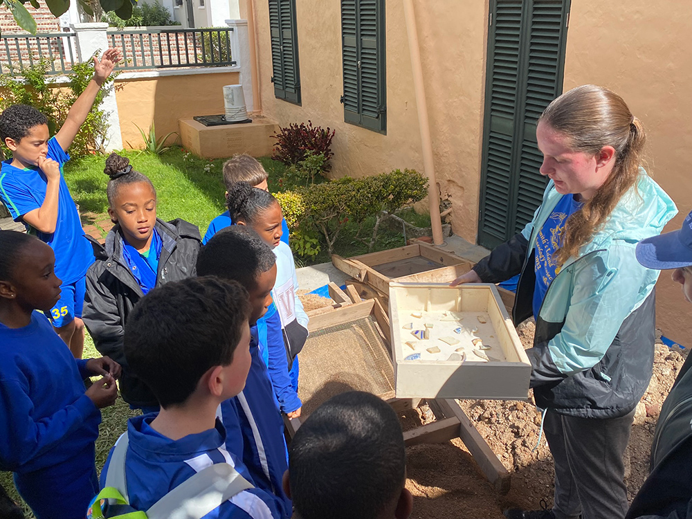 A college student holds a box full of pottery shards out for a group of school children to look at. 