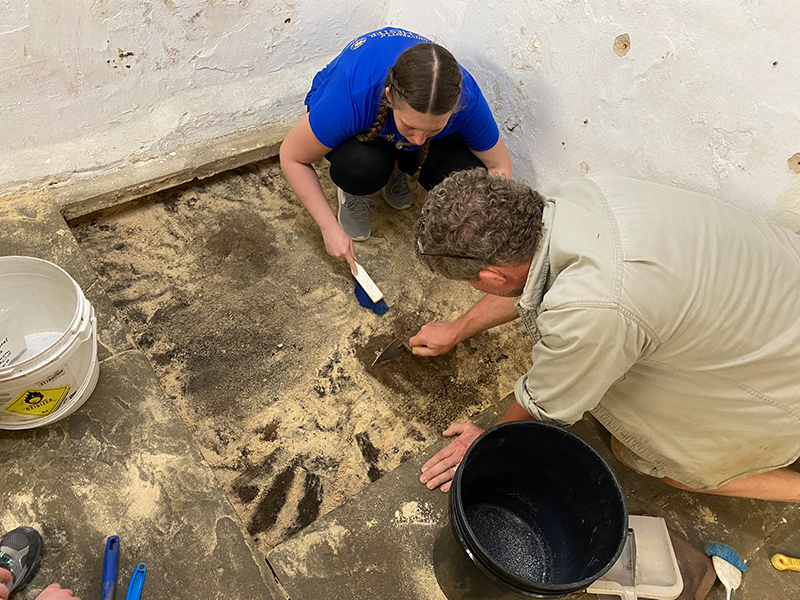 A professor and a student, kneeling on the ground inside a structure, using archaeological tools to scrape at a square of ground. 