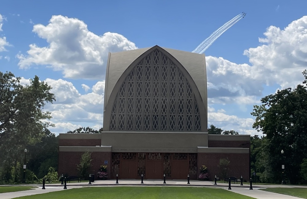 Navy fighter jets against a blue sky with white clouds over a brick building on a green lawn.