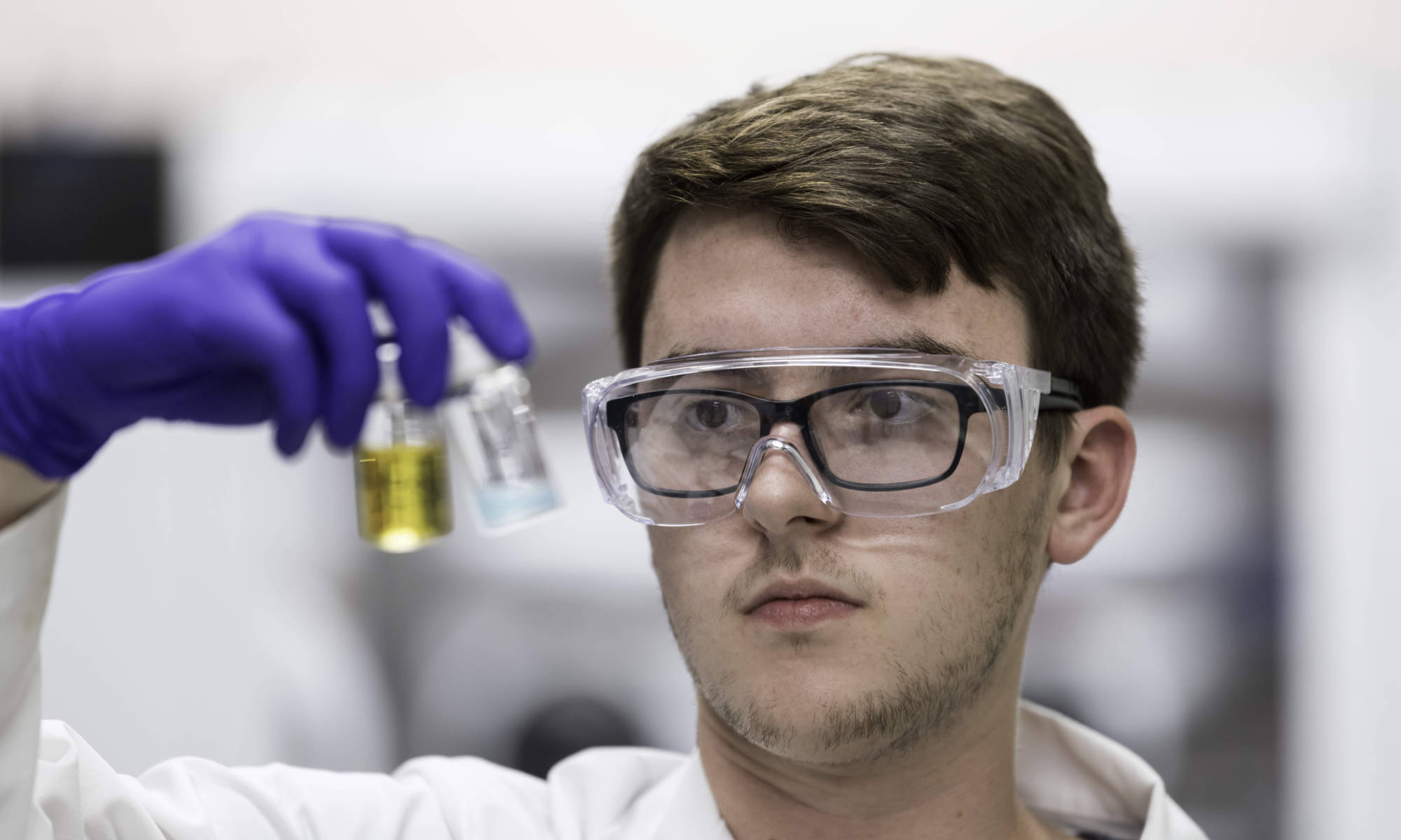 Undergraduate student in safety goggles and lab gear holds two vials of semiconducting nanoparticles with his gloved hand.