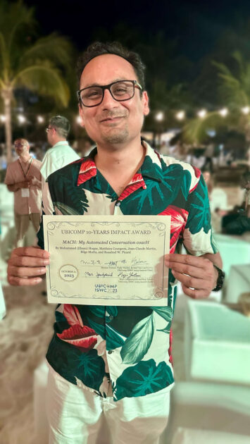 Ehsan Hoque holds his Ubicomp 10-years Impact Award certificate while smiling at the camera.