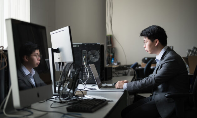 A student works at a laptop on a desk filled with computer equipment, his face is reflected in the large monitor to his left.