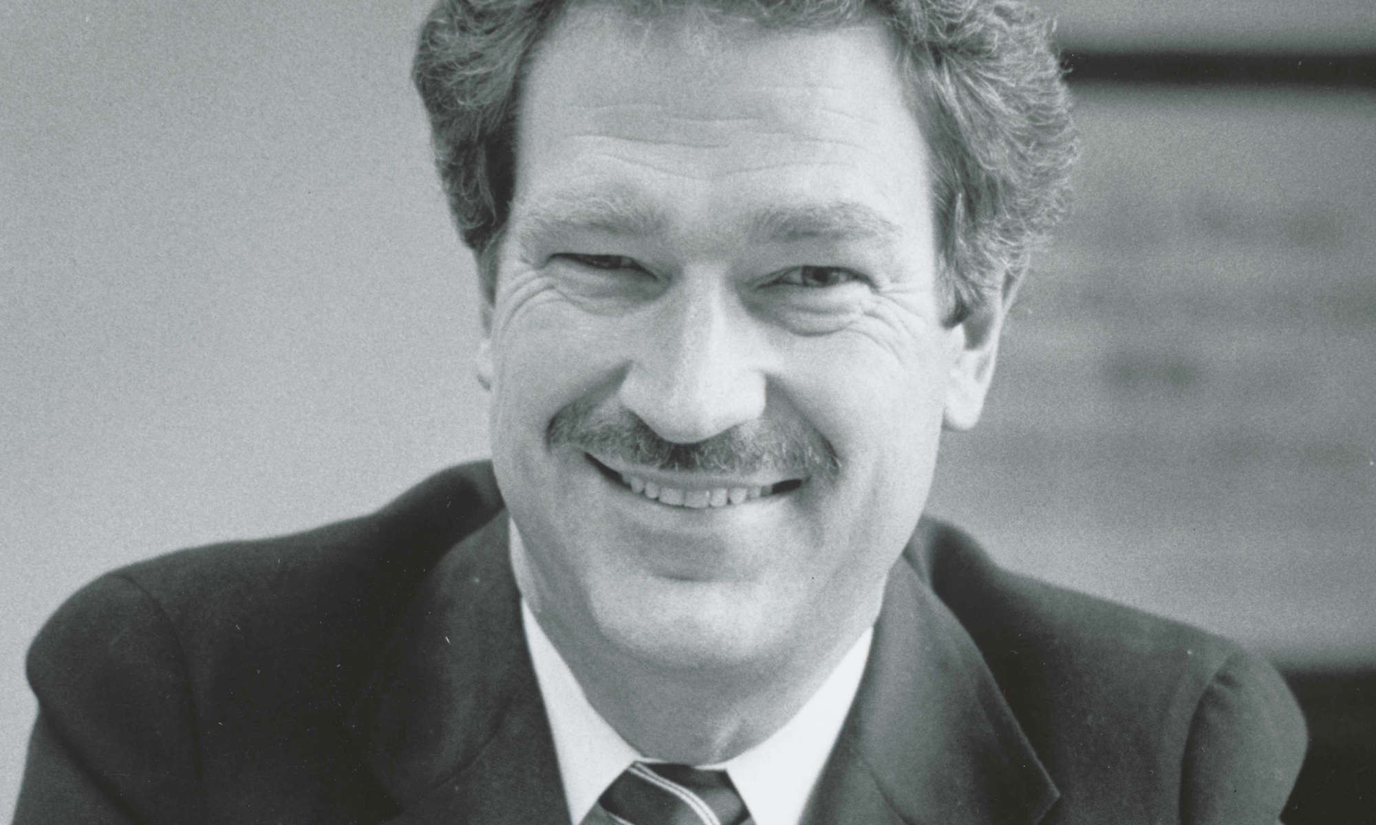 Black-and-white close up of Michael Jensen in a suit looking at the camera and smiling.