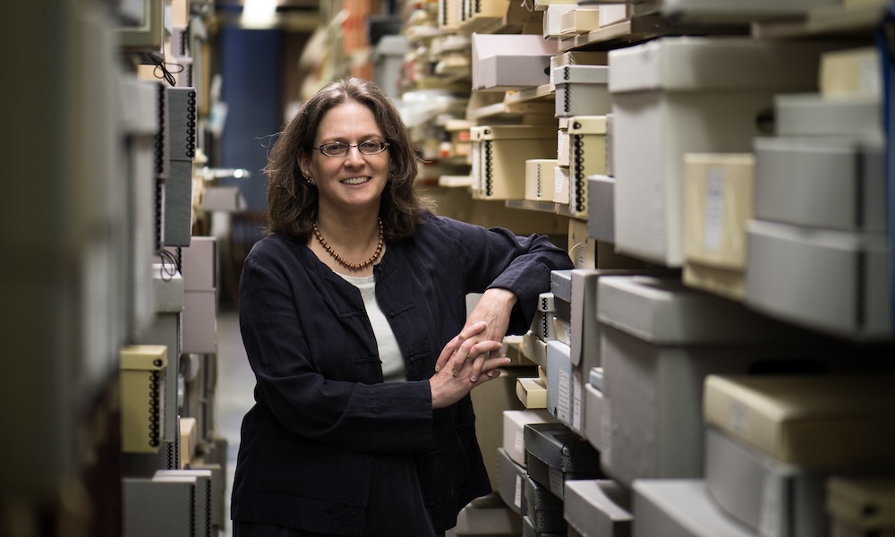 Melissa Mead smiling and leaning against shelf of gray archival boxes in archive.